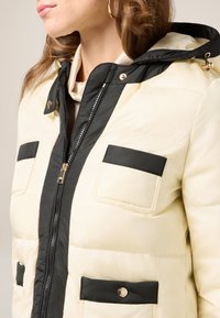 Puffer jacket in cream with black accents, featuring a zip closure, two front pockets, and a hood. Smooth texture with a quilted pattern.
