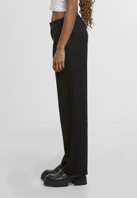 Černé kalhoty s rovnými nohavicemi vyrobené z hladké tkaniny, s bočními kapsami a skrytým předním zapínáním. Doplňují je robustní černé boty.