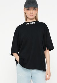 Mustelähedane oversized T-särk, mille krael on valgetes tähtedes "REPLAY", koos helesiniste kulunud teksade ja halli mütsiga.