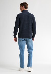 Marineren gebreide trui met een hoge kraag, gecombineerd met lichtblauwe jeans en witte sneakers, van achteren weergegeven, waardoor de gestructureerde stof zichtbaar is.