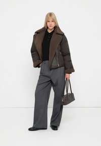 Jachetă pufătoare maro cu un guler mare din blană, purtată cu un top negru din tricot și pantaloni largi gri. Poartă o geantă neagră în mână.