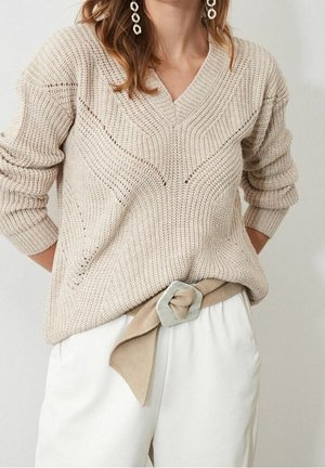 Pull en maille beige à col en V porté avec un pantalon blanc et une ceinture beige avec une grande boucle hexagonale argentée, assorti à des boucles d'oreilles pendantes.