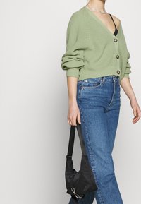 Ljusgrön stickad cardigan med upprullade ärmar och träknappar, i kombination med blå jeans och en liten svart crossbody-väska.