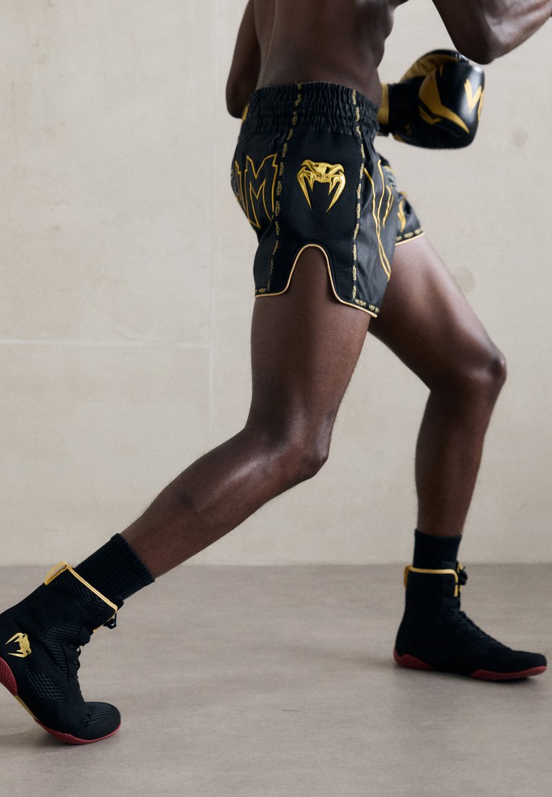 Black boxing shorts with yellow accents and graphic logo, featuring a split hem. Black athletic boots with red soles and yellow straps.