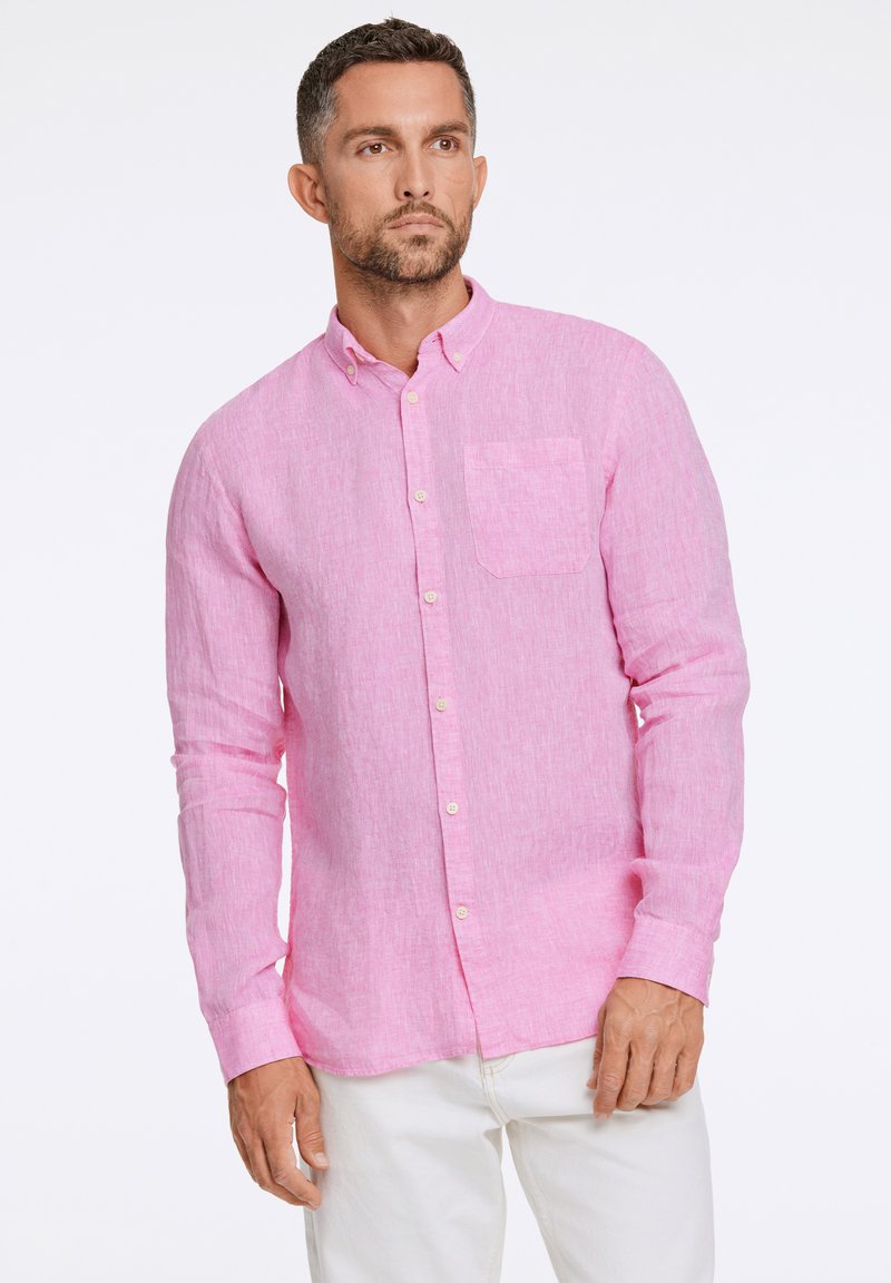 Camicia in lino rosa chiaro a maniche lunghe con colletto a punta, taschino sul petto e bottoni bianchi. Indossata con pantaloni bianchi.