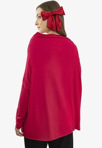 Kobieta zwrócona w lewo, ubrana w jasnoczerwony szal i dużą czerwoną kokardę we włosach na jednolitym białym tle.