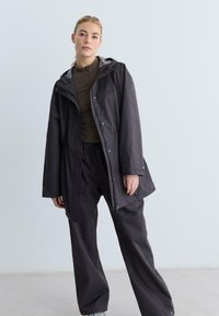 Frau in lockerer schwarzer Jacke und Hose mit grünem Reißverschlussoberteil, die vor einer schlichten hellgrauen Wand steht.