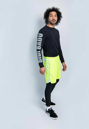 Top nero a maniche lunghe con scritte grigie, abbinato a pantaloncini giallo brillante. La modella indossa leggings neri e scarpe da ginnastica. Design semplice e sportivo.
