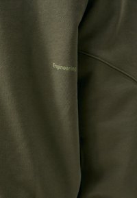 Olivenfarvet sweatshirt med ribbet tekstur og ordet "Engineering" i lys grøn. Har pæne sømme og et moderne design.