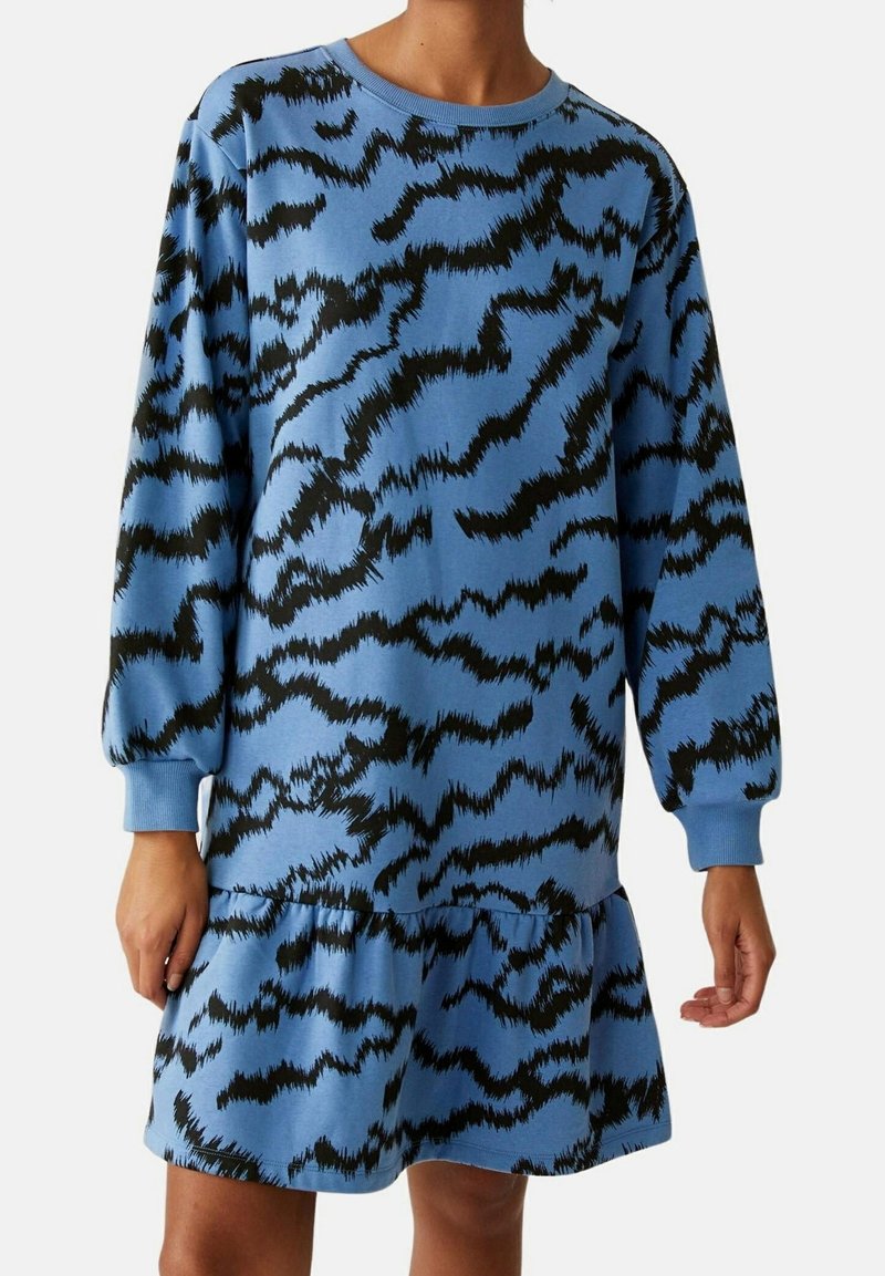 Robe bleue à manches longues avec des motifs abstraits en zigzag noirs et un ourlet à volants, portée par une personne debout.