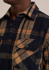 Marineblauwe en beige geruite flanellen shirt met knoopsluiting aan de voorkant, een opgestikt zakje en kraag. De stof heeft een zachte textuur met zichtbare weefdetails.