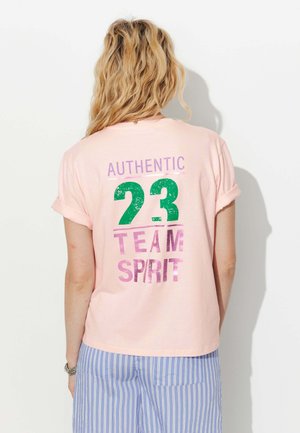 T-shirt en coton rose clair avec un texte audacieux en vert et rose indiquant "AUTHENTIC 23 TEAM SPIRIT" au dos, avec des manches roulées.