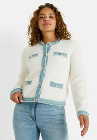 Cárdigan blanco texturizado con ribete azul y solapas de bolsillo a juego, con botones circulares. Modelo lo lleva con jeans de color azul claro.