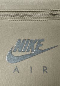 Detailní záběr šedého loga Nike Air tištěného na béžové tkanině pod švem zipu.