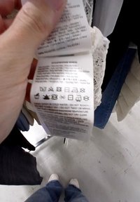 Etichetta con istruzioni per la cura e icone stampate in nero su un'etichetta di tessuto bianca, decorata con dettagli in pizzo lungo il bordo superiore.