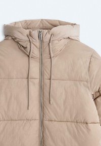Beżowa kurtka pikowana z kapturem, zamkiem błyskawicznym z przodu, sznurkami ściągającymi i poziomymi przeszyciami na materiale. Lekka tkanina.