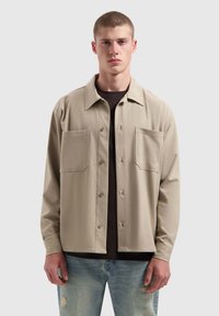 Lichtbeige long-sleeve shirt met een kraag, twee voorzakken en knoopsluiting, gelaagd over een donker t-shirt, gedragen met vervaagde jeans.