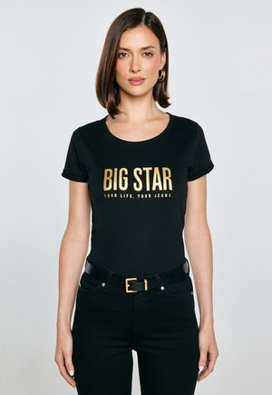 Czarny t-shirt krótki rękaw z okrągłym dekoltem z złotym napisem: "BIG STAR YOUR LIFE. YOUR JEANS." Noszony z czarnymi dżinsami i paskiem.