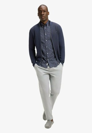 Homme portant un pull à fermeture éclair bleu marine sur une chemise bleue à motifs boutonnée, pantalon gris clair, et chaussures grises sans lacets, posant avec les mains dans les poches.