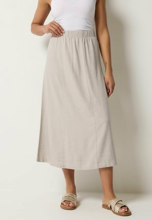 Femme portant une jupe midi beige avec une taille élastique, un haut blanc sans manches et des sandales tissées à enfiler, debout contre un mur uni.