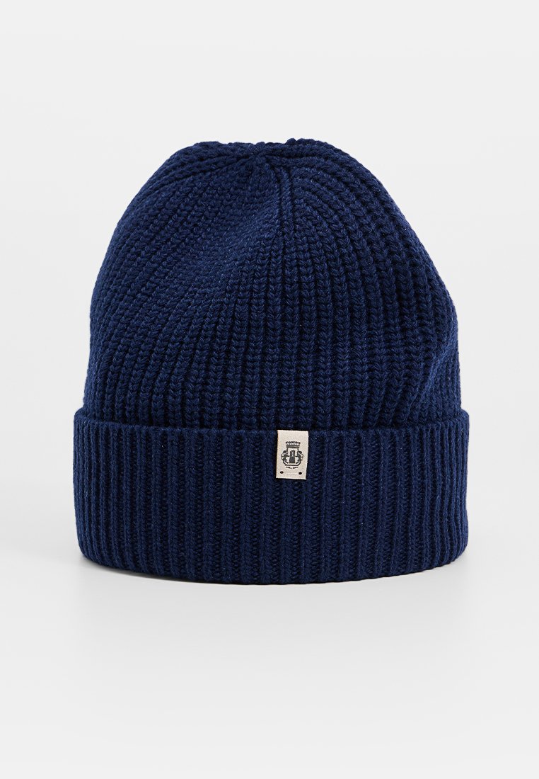 Granatowo-niebieska czapka dzianinowa z prążkowanego materiału, z podwiniętym mankietem i małą prostokątną beżową łatką logo na środkowej części przodu.