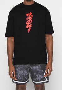 Hombre que lleva una camiseta negra con un gráfico rojo de "NO1" y pantalones cortos deportivos grises de diseño con ribetes blancos.