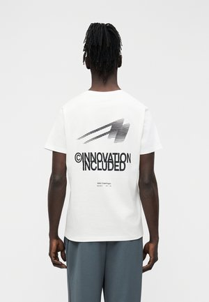 Hombre con rastas de espaldas, vistiendo una camiseta blanca con texto gráfico negro que dice "INNOVATION INCLUDED" y pantalones grises.