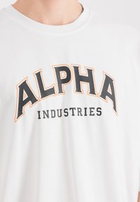 Biela bavlnená tričko s veľkým čiernym a oranžovým potlačou "ALPHA" nad "INDUSTRIES." Klasický dizajn s okrúhlym výstrihom. Jednoduché a čisté usporiadanie.