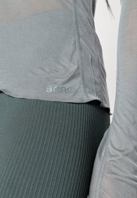 Top atlético cinza com uma textura suave e uma bainha curva, combinado com leggings verdes escuras caneladas. Apresenta um discreto logo "BORG" em azul claro.