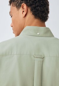 Camicia verde chiaro con colletto classico, un bottone sul retro e un dettaglio a pieghe. Realizzata in tessuto liscio con una texture morbida.