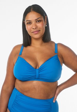 Enesetunud naine, kellel on sirged mustad juuksed, seljas ere sinine bikiin, naeratab käsi puusal valgel taustal.