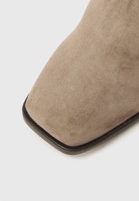 Gros plan sur une botte en daim beige avec un bout carré et un bord de semelle noir sur un fond clair.