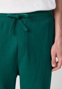 Teal joggebukser har et strukturert stoff, elastisk midjebånd med snor og sidelommer. Nærbilde av midjebåndet og håndplassering.