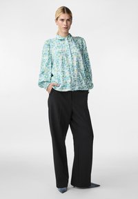 Blusa blu chiaro con un motivo colorato, dotata di colletto e maniche lunghe, abbinata a pantaloni neri a gamba larga e scarpe blu a punta.