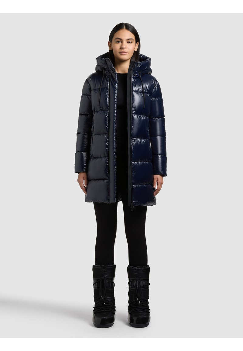 Sieviete, kas ģērbta tumši zilu spīdīgu dūnu mēteļu ar kapuci, melnās legingās un melnās ziemas zābakos, stāvot pret vienkāršu pelēku fonu.