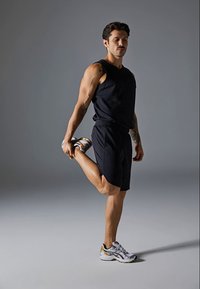 Mann i svart tanktop og shorts strekker quadriceps ved å holde en fot bak seg mot en grå bakgrunn.