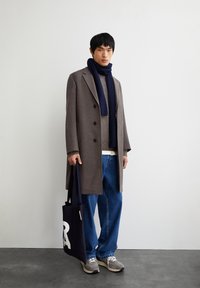 Grauer Wollmantel mit doppelt geknöpftem Verschluss, navyblauer Rippenschal, beigefarbener Strickpullover, blaue weite Jeans und graue Sneakers. Schwarze Tote-Tasche mit weißen Buchstaben.