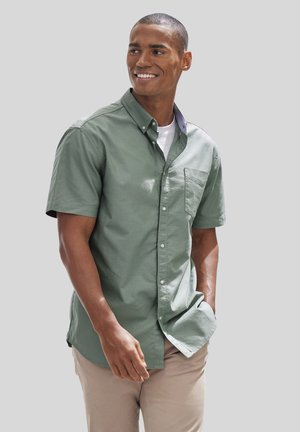 Short-sleeve button-up shirt in olive green cotton with a chest pocket and a contrasting striped collar. Worn with light beige pants.