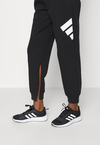 Černé joggingové kalhoty s bílými páskami a postranními rozparky, doplněné černými sportovními botami s bílými pruhy a texturovaným svrškem.
