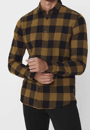 Uomo che indossa una camicia a maniche lunghe a quadri giallo senape e nero, che stringe un polso con l'altra mano, mostrando un anello.