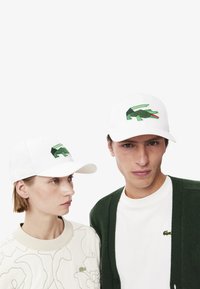 Gorra de béisbol blanca con un logo de cocodrilo verde. La tela está estructurada con una visera curva, llevada por dos modelos en atuendo casual.