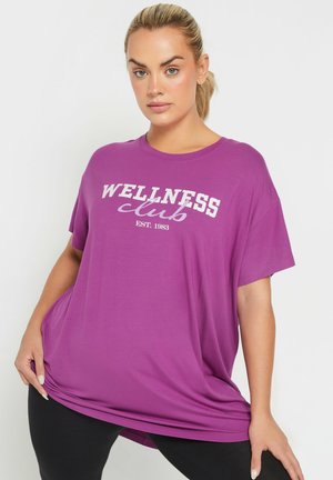 Kobieta o blond włosach związanych w kucyk, ubrana w luźny fioletowy T-shirt z napisem "WELLNESS club EST. 1983" oraz czarne legginsy na białym tle.
