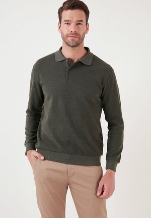 Suéter de polo de manga larga en verde oscuro, con cuello tipo polo, tres botones y puños y dobladillo acanalados, combinado con pantalones beige claros.