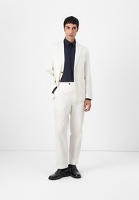 Witte suit met een op maat gemaakte pasvorm, voorzien van een enkele knoopsluiting en zijzakken. Gecombineerd met een donkerblauw overhemd en zwarte dresschoenen.