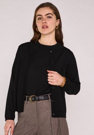 Femme portant un cardigan noir sur un haut noir, un pantalon marron taille haute à plis, une ceinture noire, des boucles d'oreilles créoles dorées et un bracelet manchette en bois.