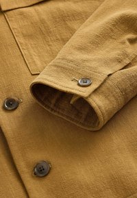 Camisa marrón con textura, con puño vuelto y dos botones oscuros. Presenta un bolsillo en el pecho y un cuello recto.