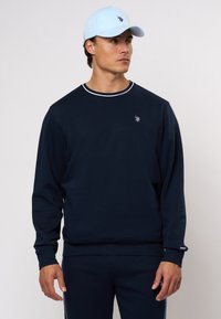 Tamsiai mėlynas sportinis džemperis su baltu apykaklės apvadu, ant krūtinės yra mažas logotipas. Suderintas su derančiomis tamsiai mėlynomis kelnėmis. Šviesiai mėlyna kepurė.