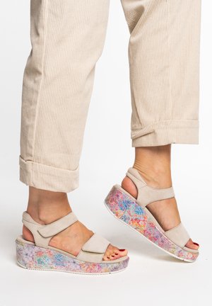 Feet wearing beige open-toe platform sandals with colorful floral-patterned soles and beige corduroy pants with rolled cuffs.