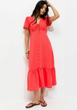 Mujer con vestido midi coral abotonado, de manga corta y bajo con volantes, complementado con pulseras y sandalias marrones.