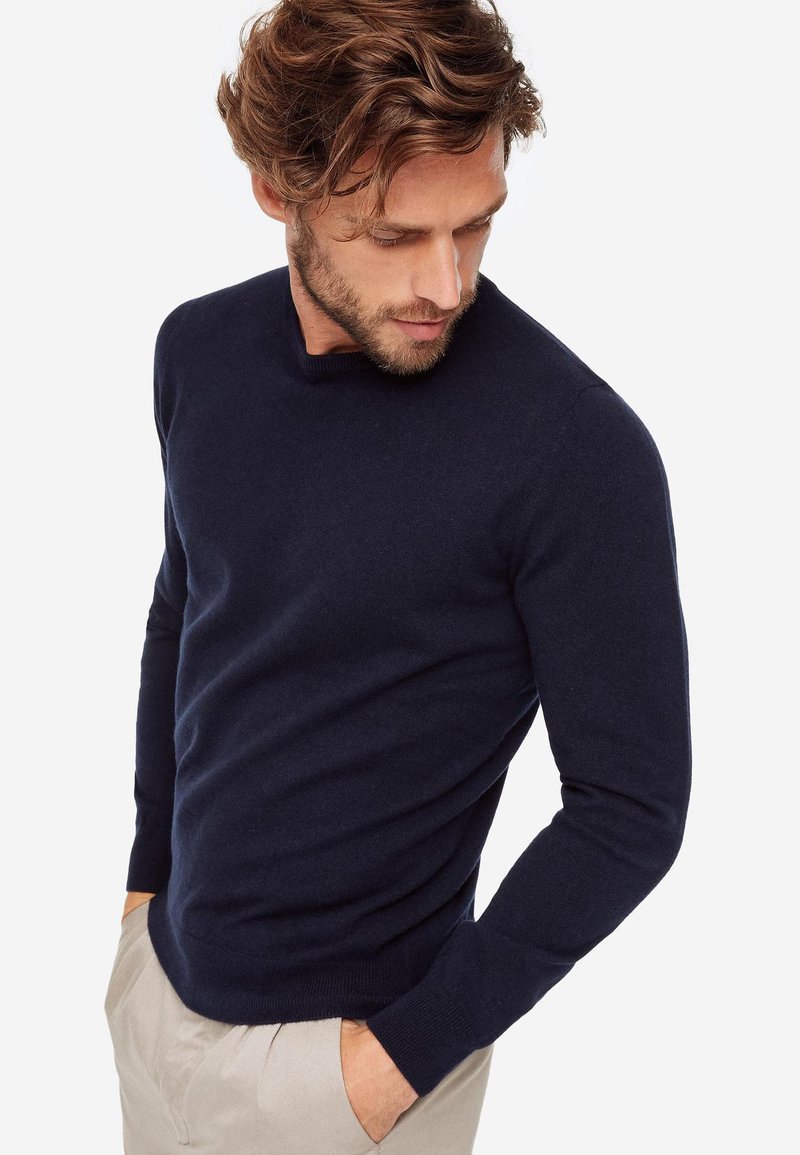 Pull en maille bleu marine avec un col rond et des manches longues, présentant une texture lisse et une coupe ajustée, associé à un pantalon beige.